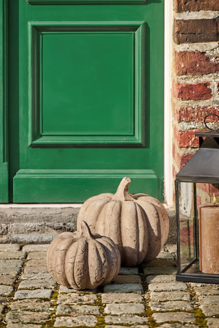 Two natural terracotta pumpkins in different sizes on a cobblestone doorstep beside a green front door and a lit lantern candle