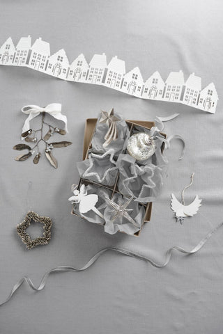 Assorted silver and white Christmas ornaments including stars, angels, a mercury glass bauble, and ribbon, arranged in a box beside a paper house garland