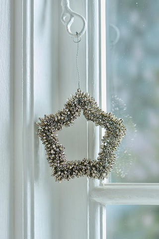 Silver beaded star ornament hanging from a white wall hook against a window