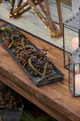 Weathered grey metal tray holding dried juniper berry garlands on a rustic wooden table beside star-cut lanterns with lit candles