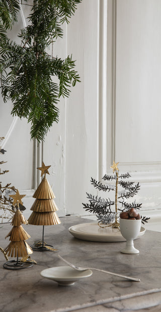 Decorative table setting with gold Christmas trees, greenery, and a white candle on a marble surface.